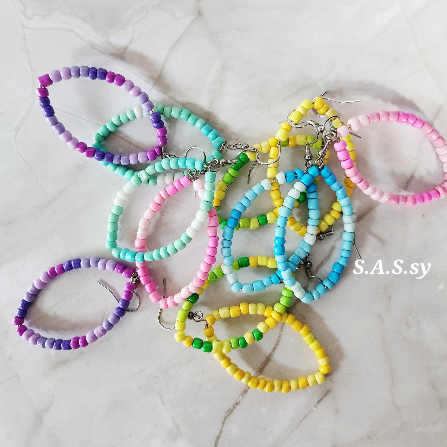 Colorful beaded hoop earrings on a marble surface with 'S.A.S.sy' branding.