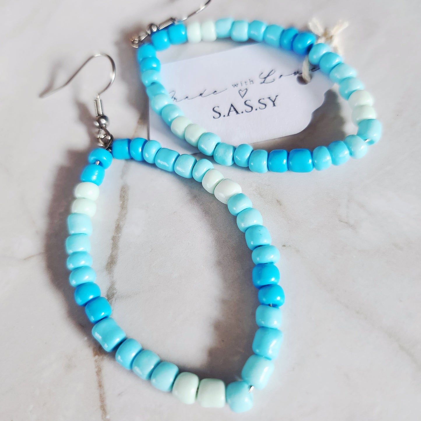 Aqua beaded hoop earrings with S.A.S.sy brand name on marble background