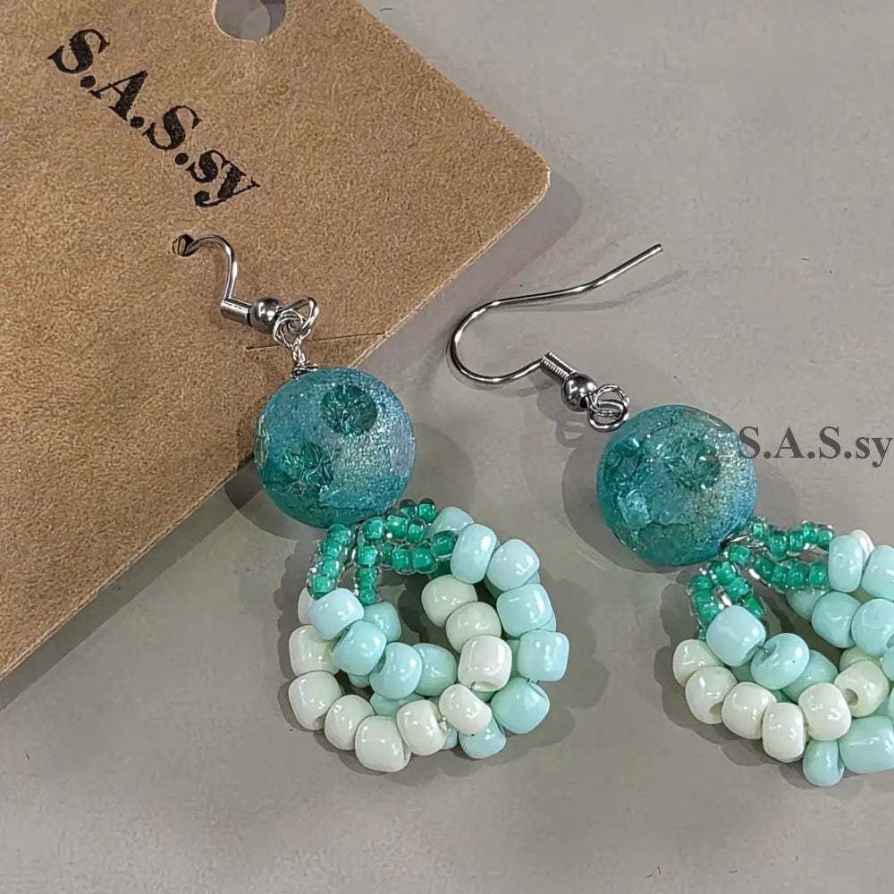 Beaded earrings with blue and green beads on a gray surface, accompanied by a branded card.