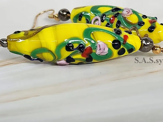 A pair of yellow dangle earrings with blue and red polka dots and silver accents, displayed against a marble background.