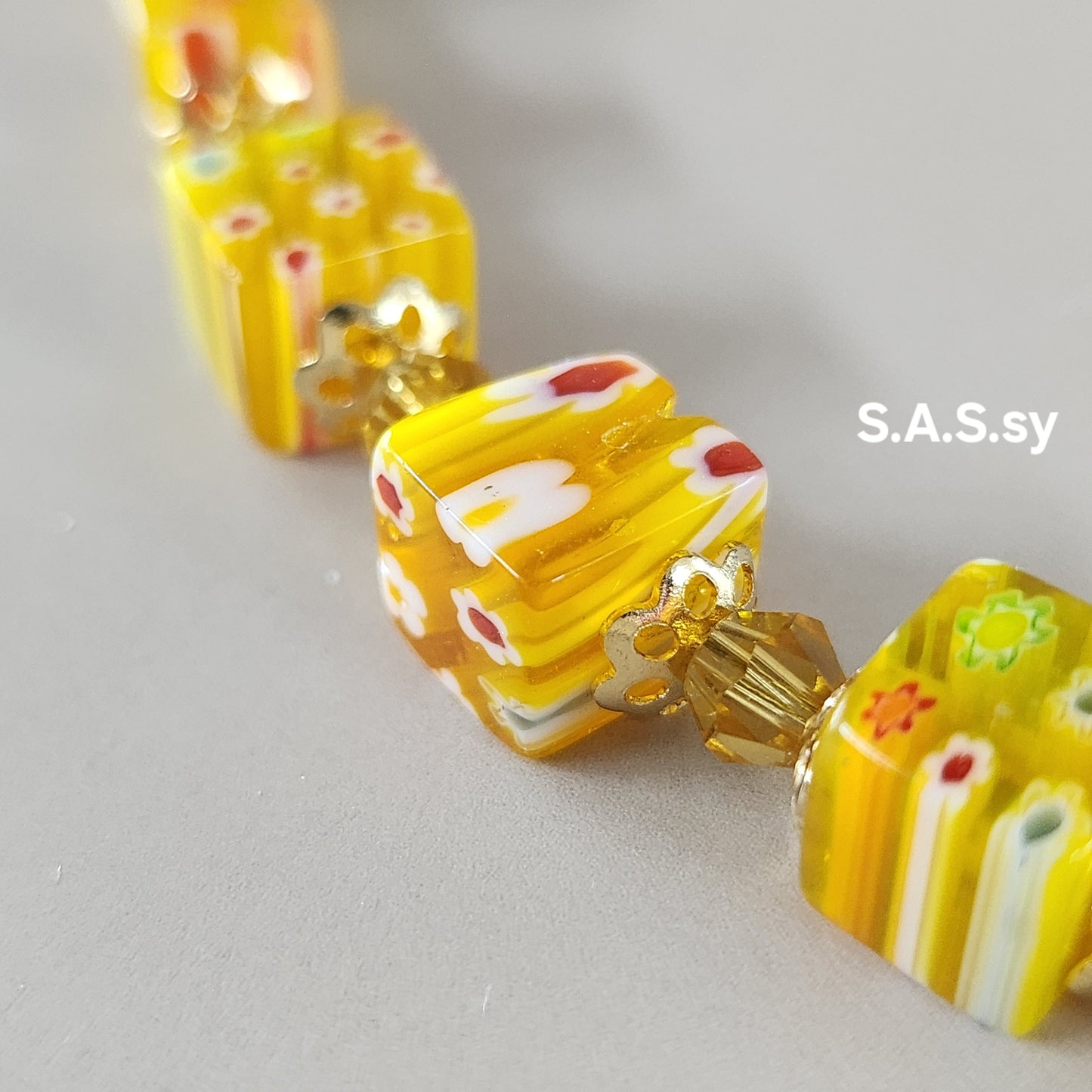Close-up of a yellow beaded necklace with floral patterns on a light gray background, branded 'S.A.S.sy'.
