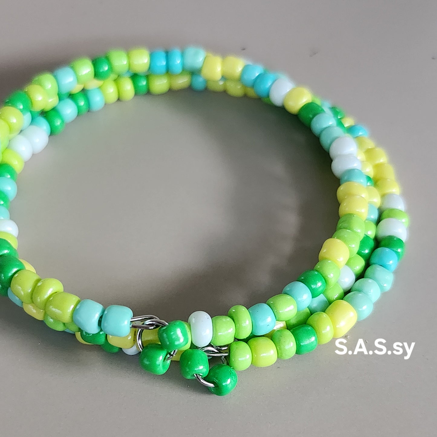 Green, yellow beaded  coil bracelet on a gray background with 'S.A.S.sy' branding.