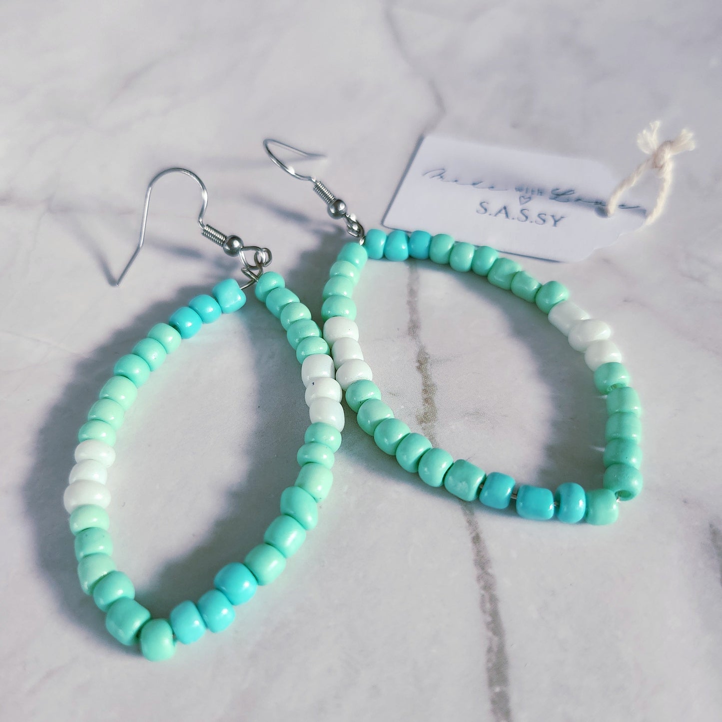 Turquoise and white beaded hoop earrings on a marble surface with 'SASSY' branding.