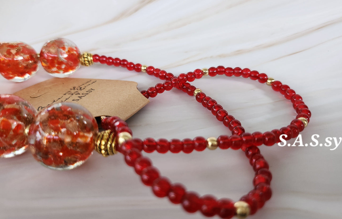 Red beaded necklace with gold accents on a light background, featuring the brand 'S.A.S.sy'.