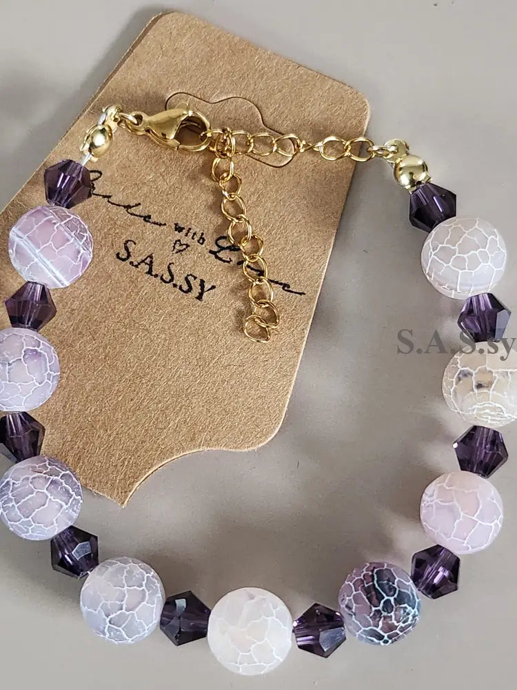 Bracelet with purple and white beads on a beige card with 'SASSY' branding.