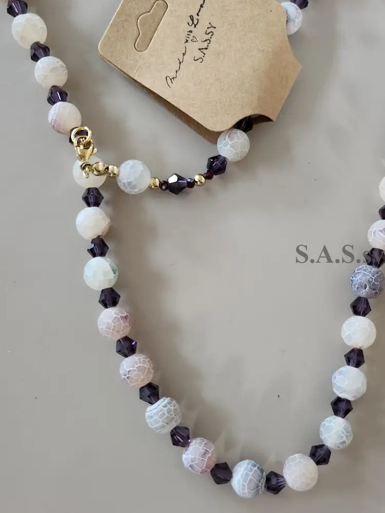 Necklace with white and purple gemstone beads on a gray surface, featuring a brand tag.