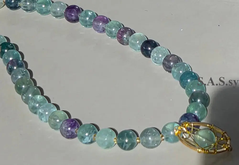 A beaded necklace made of polished round gemstone beads in shades of light blue, teal, and purple, with a gold-toned geometric cage pendant, displayed on a light gray surface.