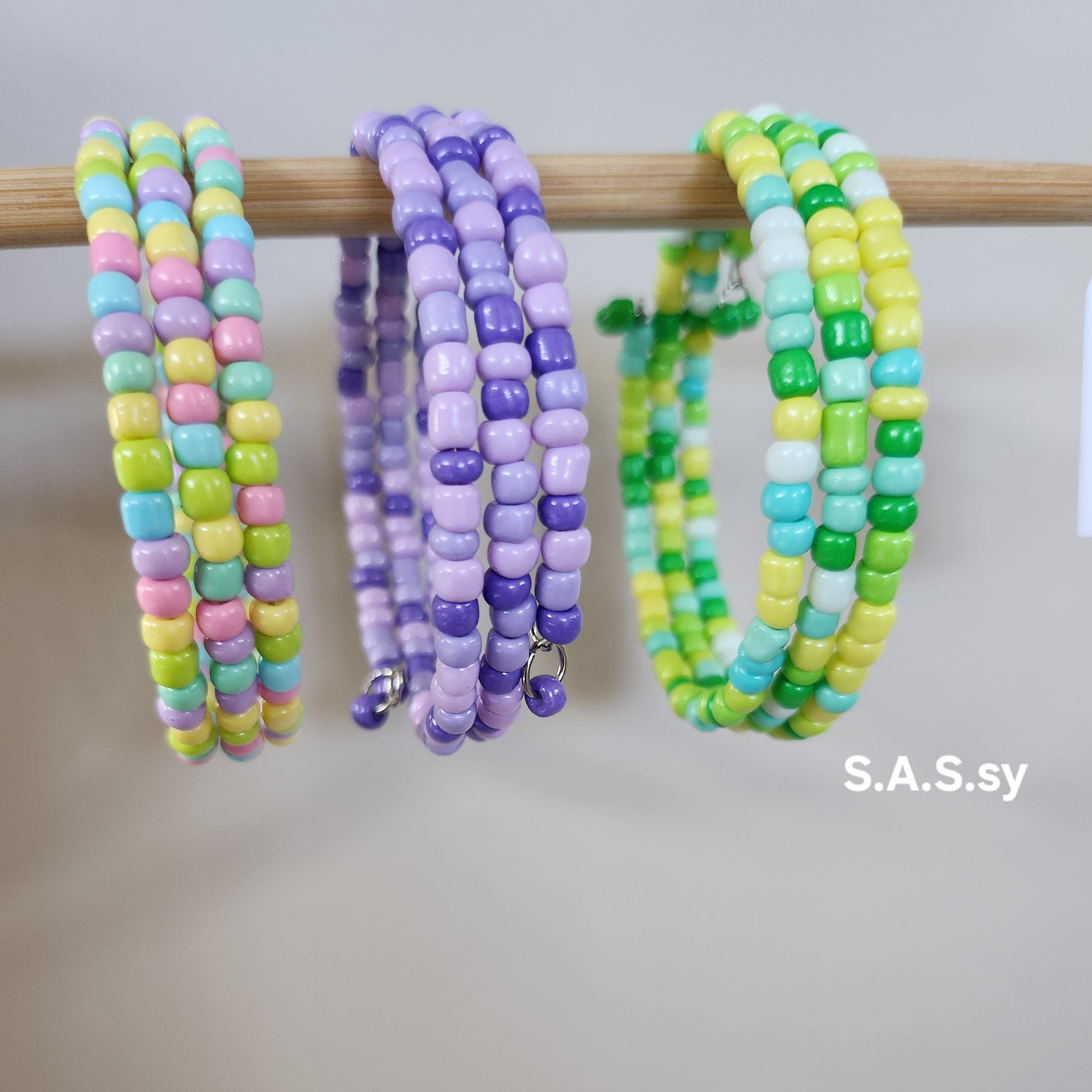Colorful beaded bracelets on a wooden stick with 'S.A.S.sy' branding.