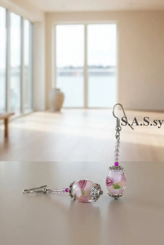 A pair of handmade dangle earrings featuring translucent pink glass beads with swirl patterns, silver filigree caps, and silver fishhook ear wires, displayed on a neutral beige background.
