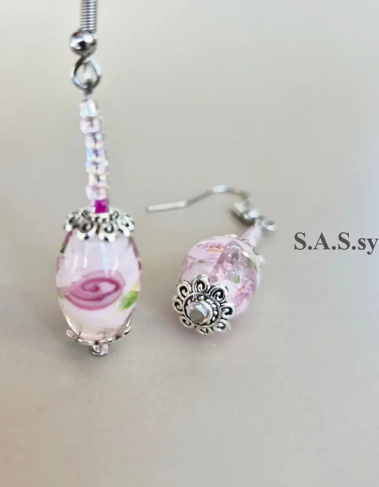 A pair of handmade drop earrings featuring pink and white floral glass beads with silver filigree caps and silver fishhook ear wires, displayed on a neutral beige background.